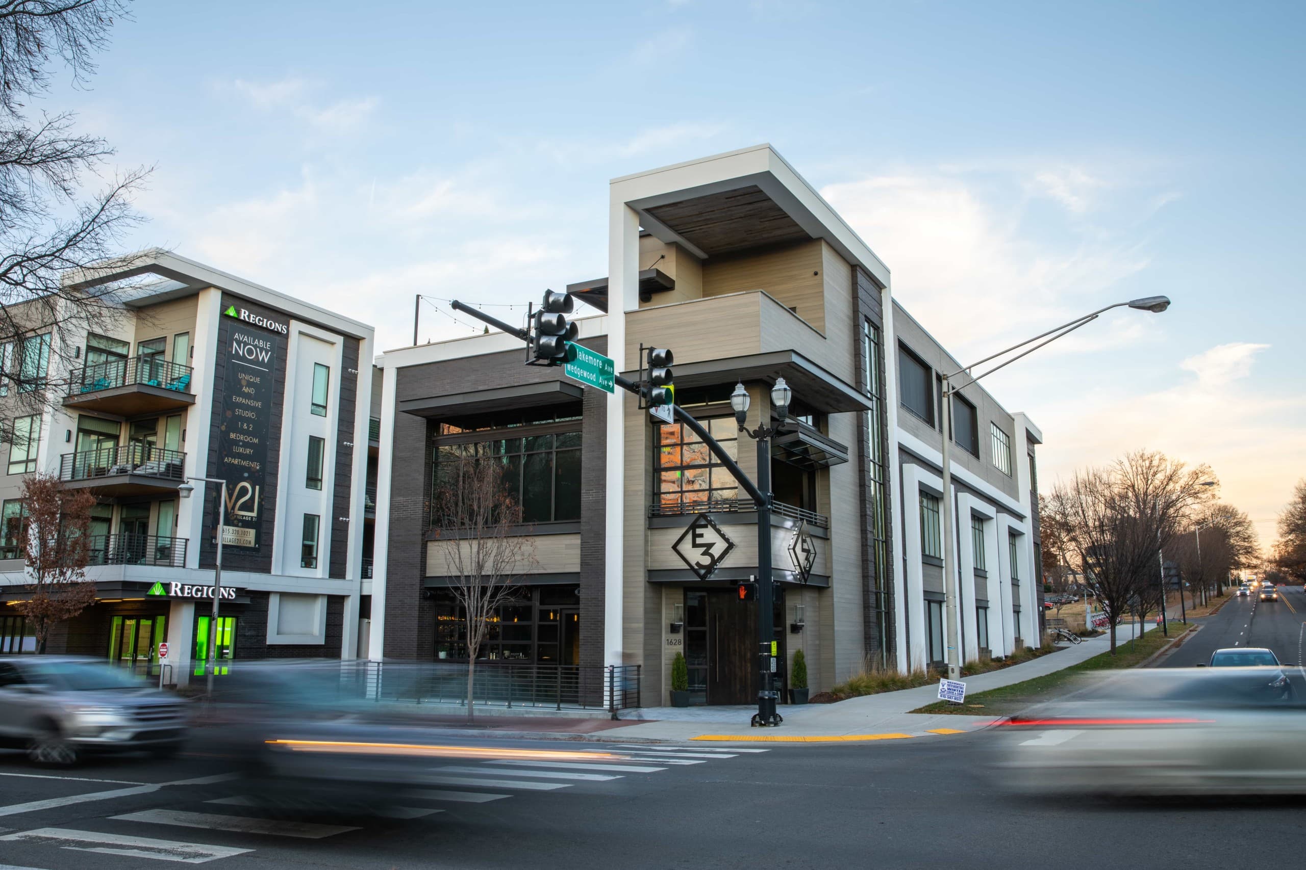 Exterior of a modern white and mud grey corner building, which has windows, and an E3 metal logo on it with planters around its entrance.