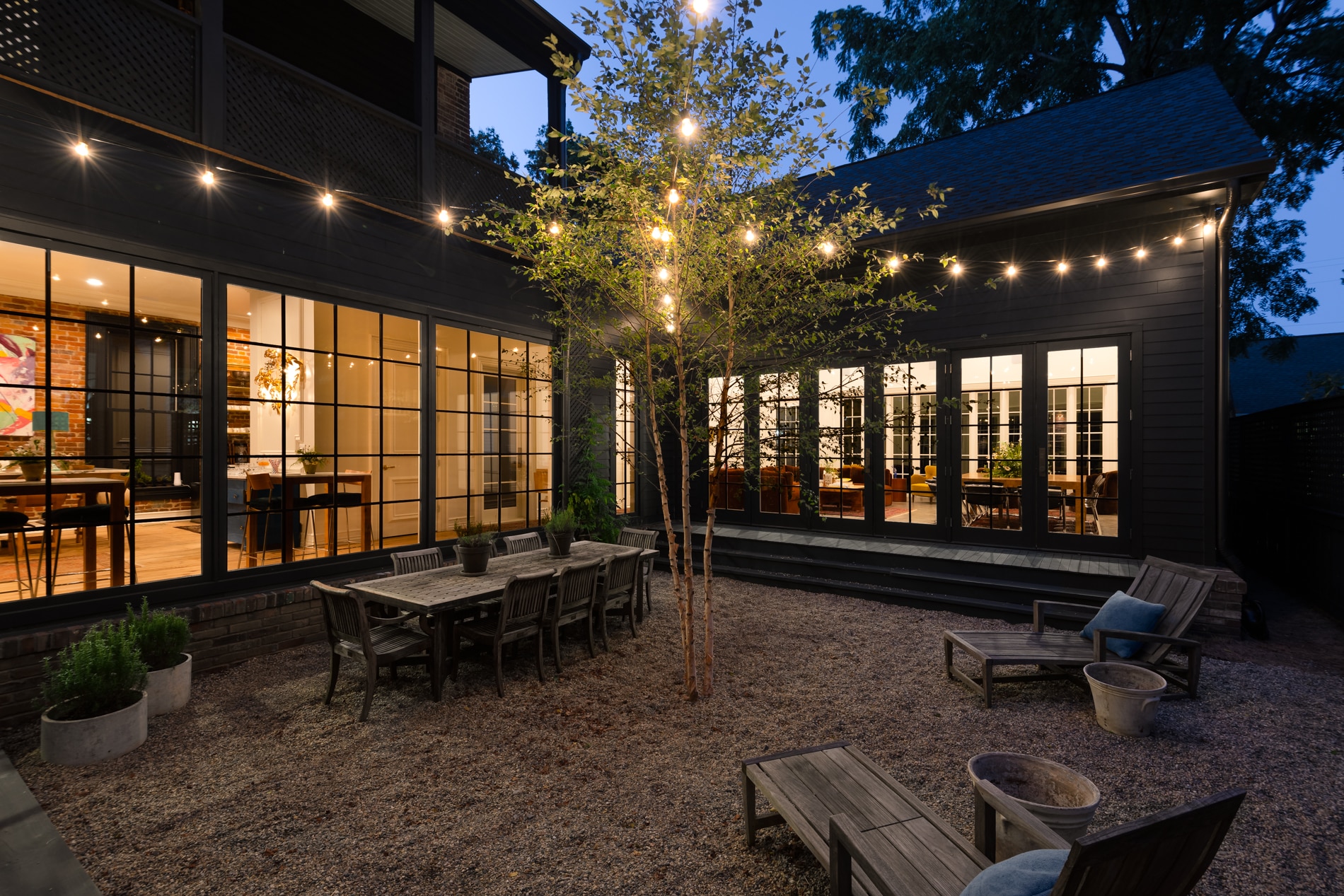 A courtyard of a historic house with a tree holding hanging lights, a wooden dining table with chairs, planters and wooden recliner chairs.
