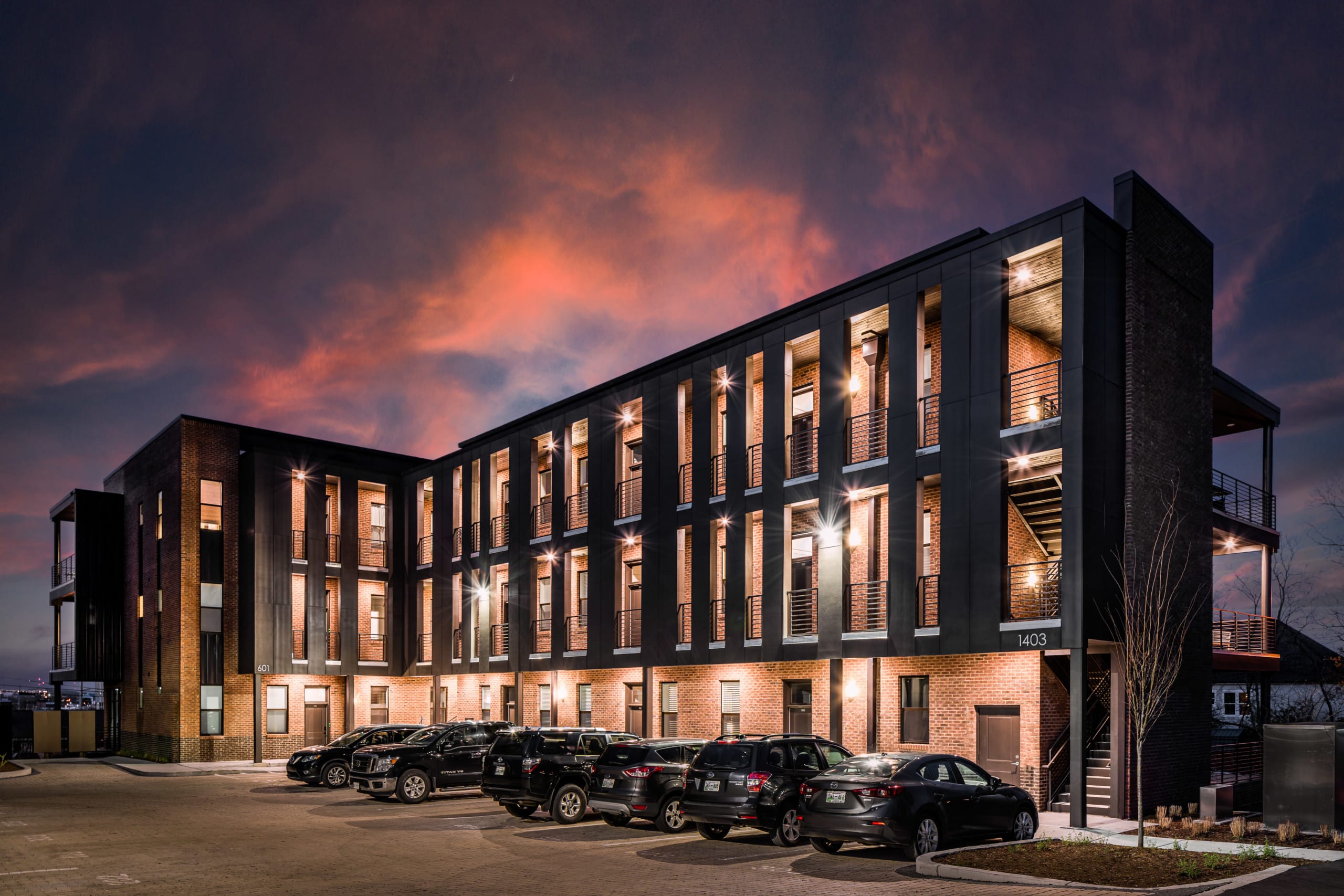A residential building in the evening, with windows, stairs, doors, and corridors. A range of cars are parked in front of the building.