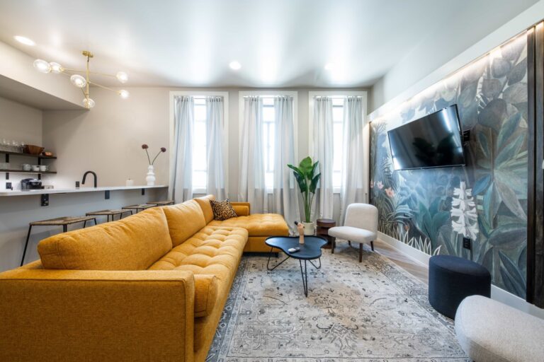 TV Lounge with white curtains, mustard sofa, tropical plants painted on a wall having TV, and open kitchen at the other side.