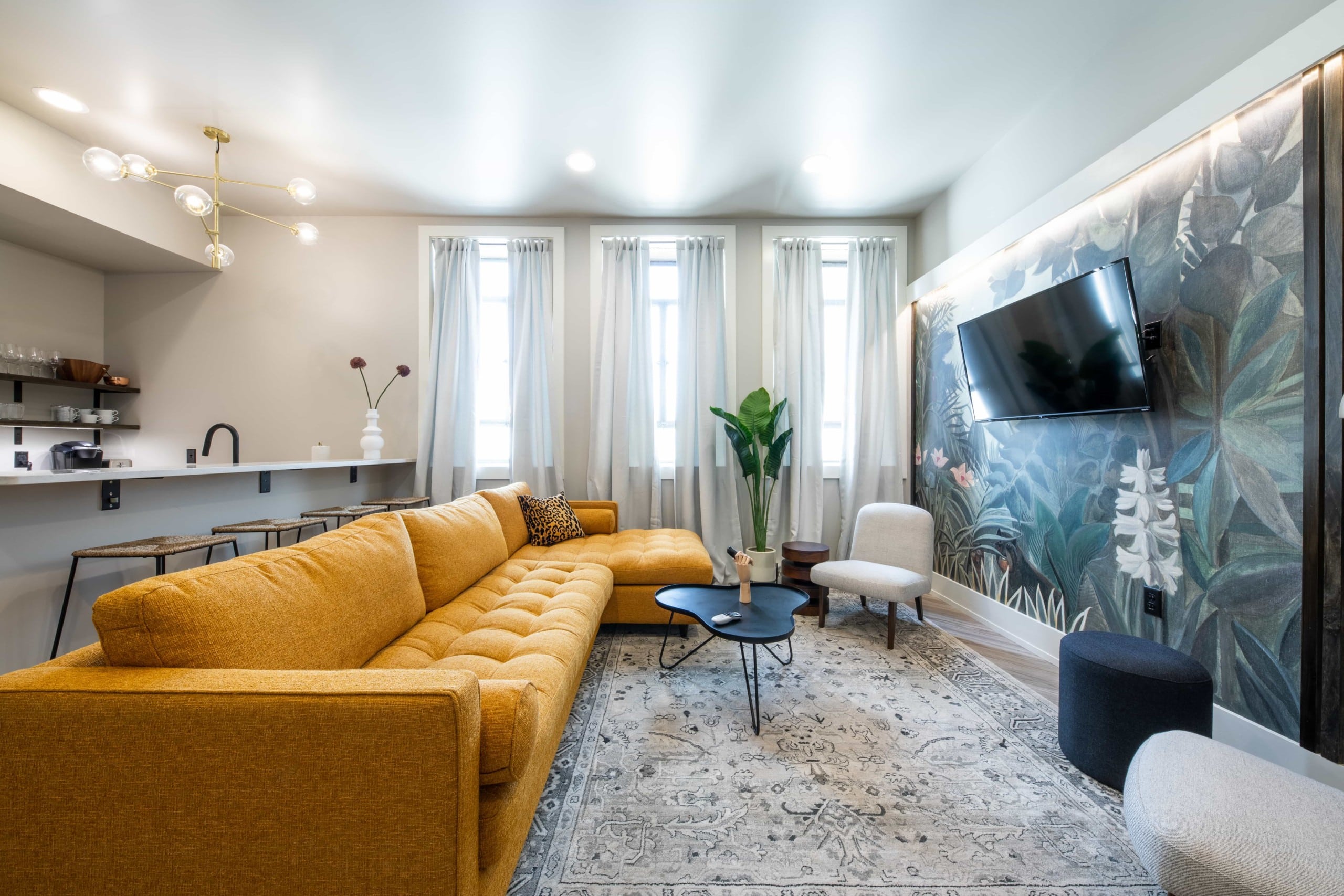 TV Lounge with white curtains, mustard sofa, tropical plants painted on a wall having TV, and open kitchen at the other side.