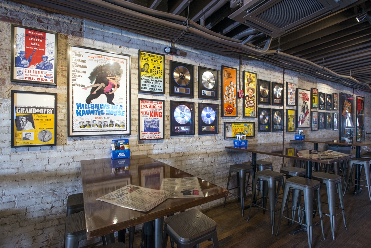 White bricked wall with 90s-themed frames, music records, theater posters, and tables with sauces in a box having "BUD LIGHT" on it.
