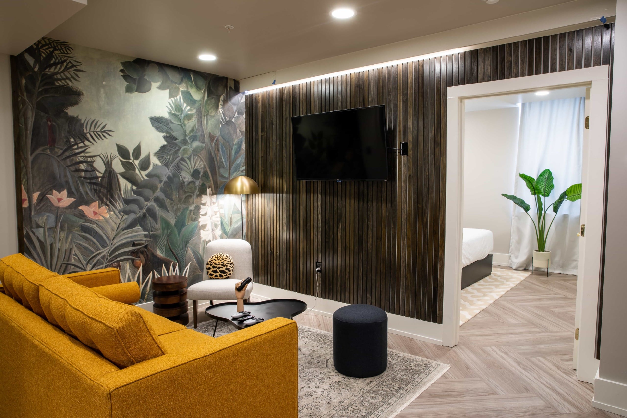 TV lounge with mustard colored sofa, wall painted with tropical leaves, a door opening to a white room having a tropical plant.