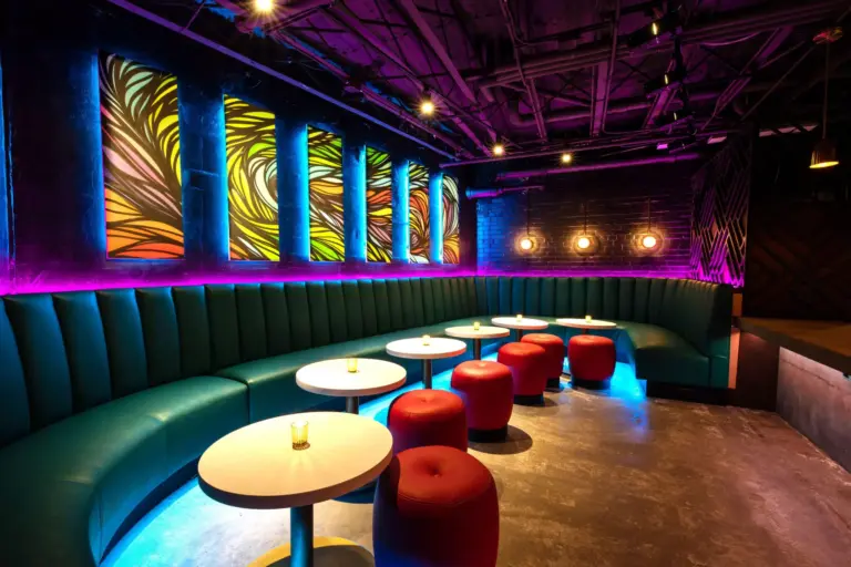 Sitting area with colorful light art on the back wall, purple lighting, teal sofa, red ottomans and circular tables candles.