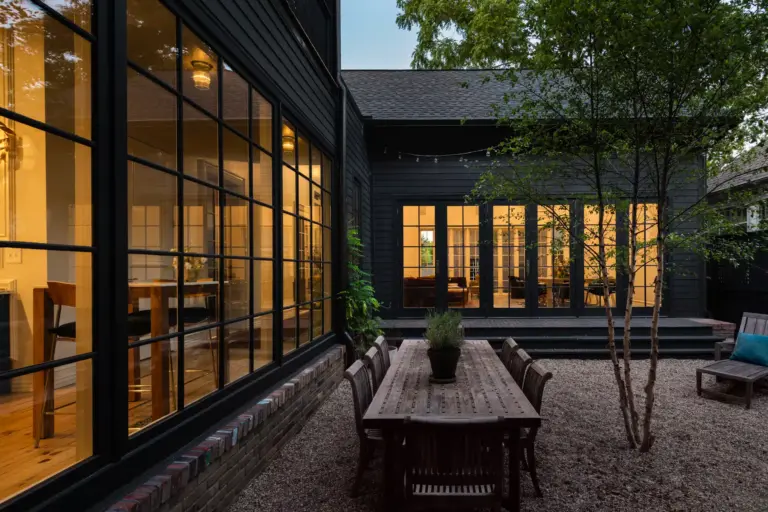 Large windows at the front and one side of a courtyard space that contains a tree, wooden dining table, chairs, and recliner chair.
