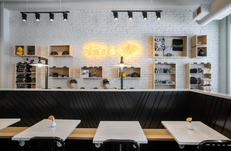 White wall with focus lights, white shelves having shirts, caps and beanie caps on display and yellow neonlight of cheese and bread.