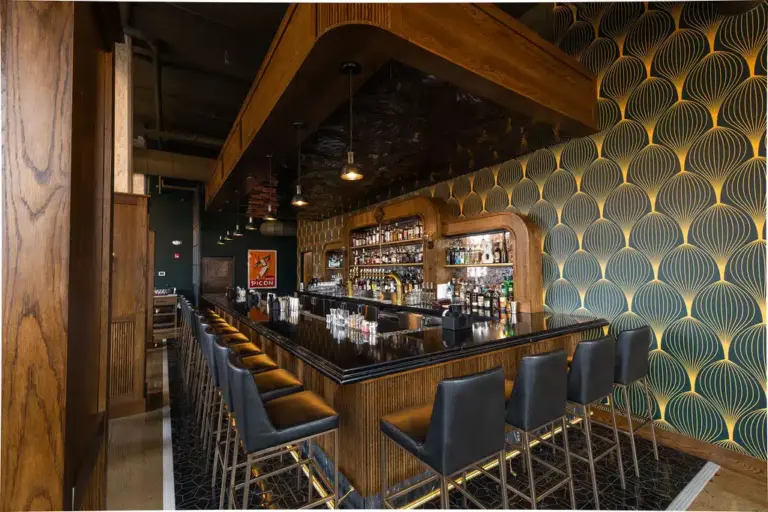 View of antique-themed bar counter with wooden shelves and black furniture, wall at the back filled with different colored bottles.