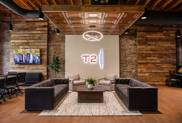 Sitting area with wooden roof, floor, black & grey sofas, and a table with a planter. The front wall has "T2" on it in orange neon lights.