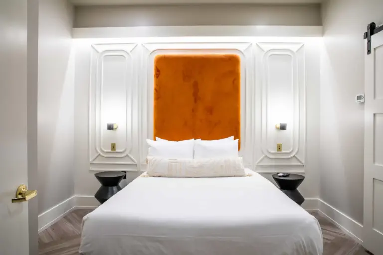 A white-themed bedroom with a rust bedhead, two black side tables, two wall lights, grey titled floor and a white bed with white pillows.