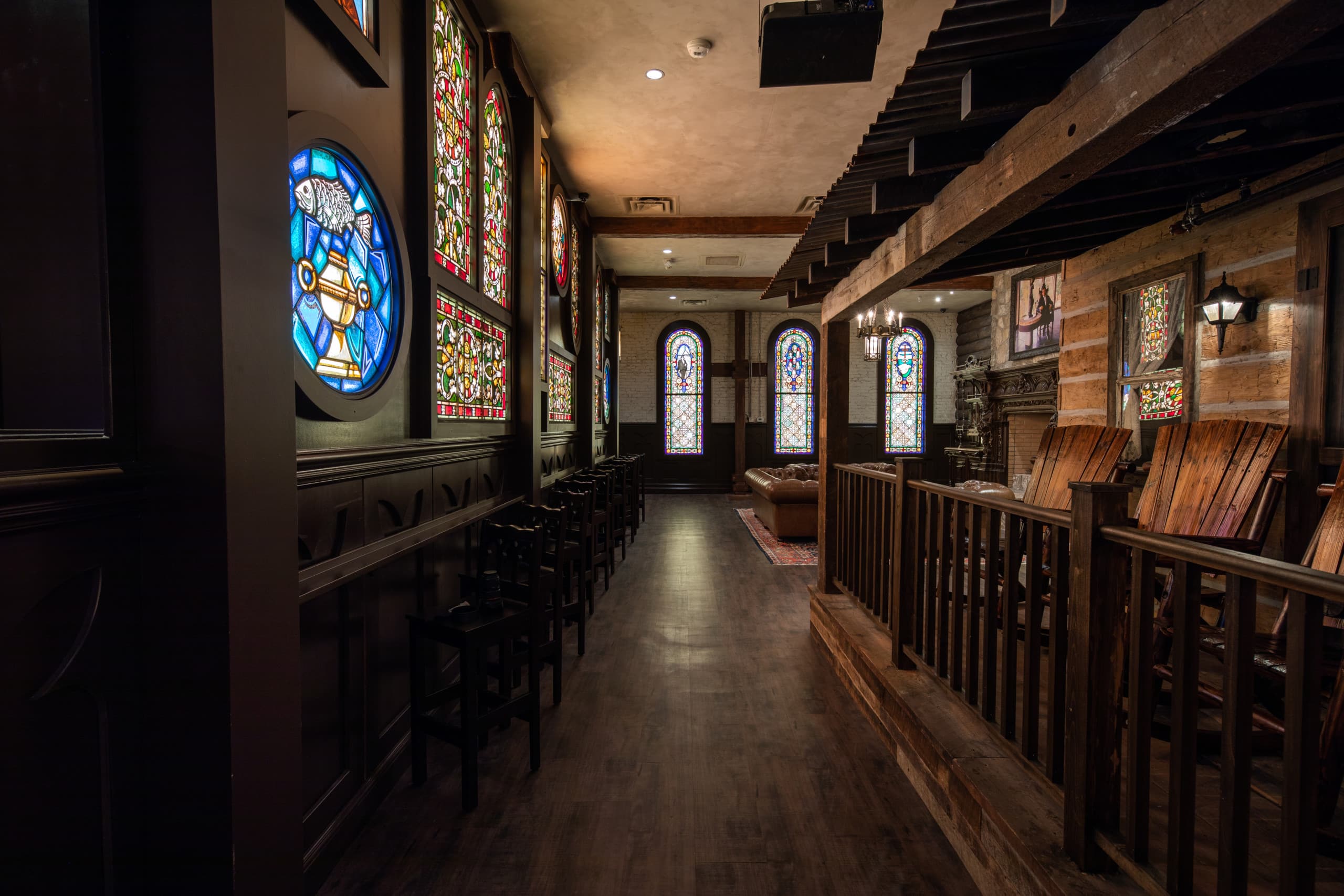 Medieval interior of a building with arched, colored glass windows having different arts, wooden sitting furniture and a painting of a man.