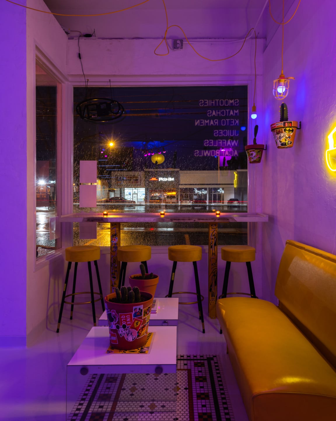 A corner of a shop with windows, white tables, yellow stools and sofas, planters on tables and walls and hanging purple lights.