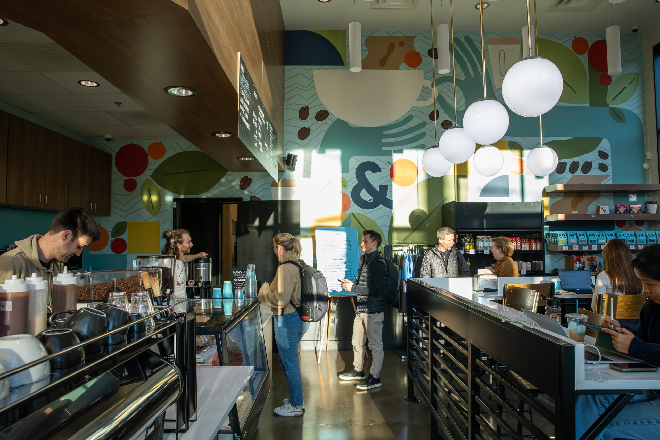 People working on laptops and having coffee, some are over the counter that has coffee beans boxes and front wall of colorful nature theme.