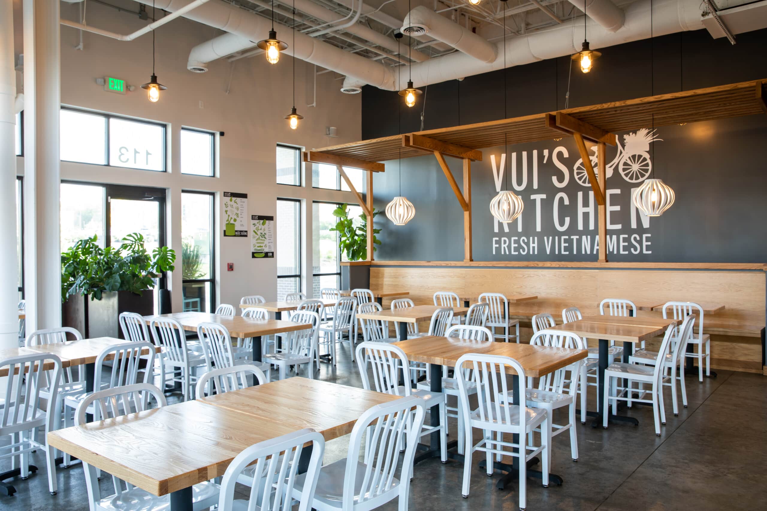 A restaurant with sitting area having wooden tables, white chairs, open kitchen and one grey wall saying "Vui's kitchen fresh vietnamese"